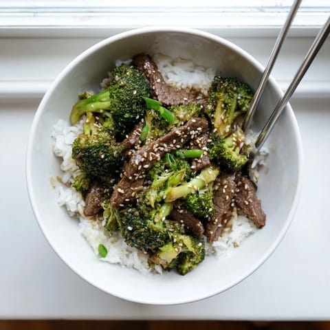 A steaming plate of Beef and Broccoli stir-fry, topped with sesame seeds and fresh green onions, ready to eat.