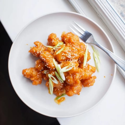 Crispy fried Orange Chicken bites coated in a sticky, glossy orange sauce, served on a white plate with chopped scallions and sesame seeds.